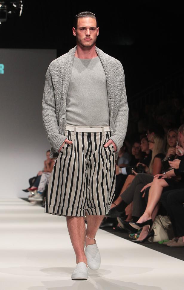 Ein Mann präsentiert Mode auf einem Laufsteg, mit gestreiften Shorts und einem grauen Cardigan.