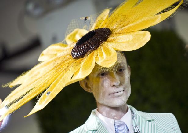 Eine Person mit weißer Gesichtsbemalung trägt einen gelben Sonnenblumenhut mit einer kleinen Spielzeugbiene.