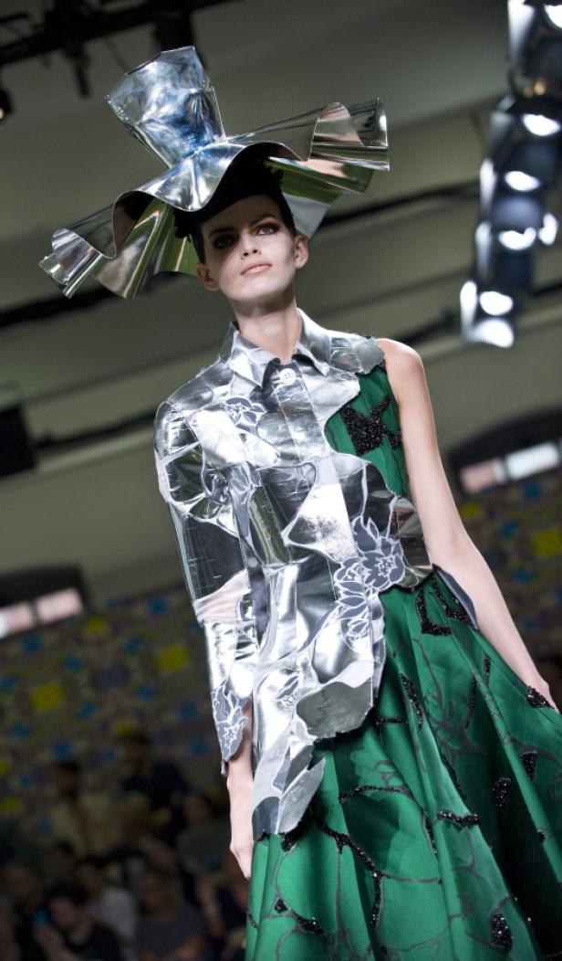 Eine Frau mit einem extravaganten Hut und einem Kleid aus glänzendem Material auf einem Laufsteg.