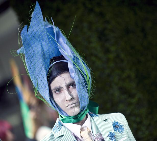 Ein Model mit blauem Hut und grün gestreiftem Anzug mit Blumenmuster präsentiert ein extravagantes Outfit.