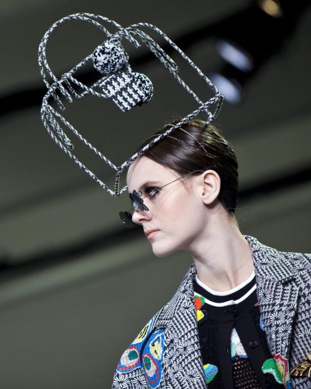 Eine Frau mit ausgefallenem Kopfschmuck und Schmetterlingsbrille auf einem Laufsteg.