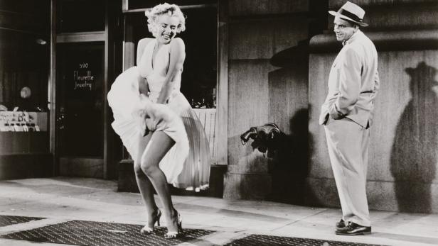 Marilyn Monroe, mit wehendem Kleid, und ein Mann stehen auf einem New Yorker Bürgersteig.