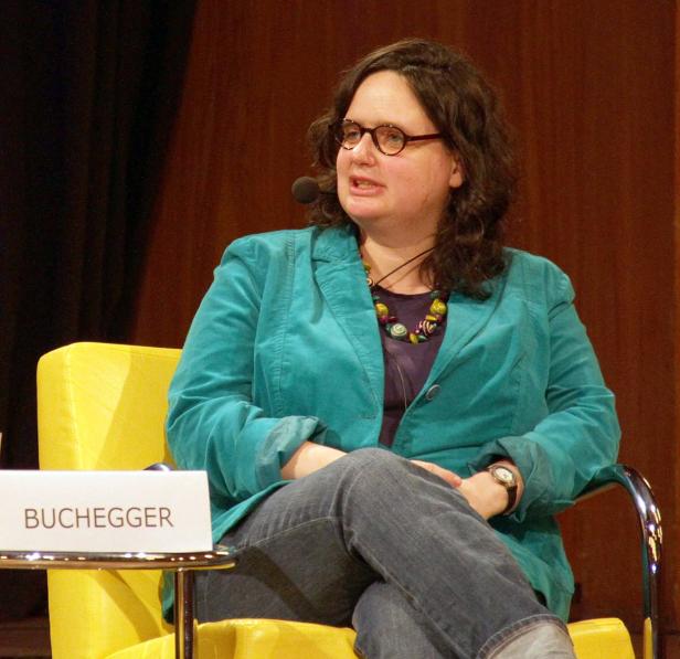Eine Frau, möglicherweise Buchegger, sitzt auf einem gelben Stuhl und spricht.