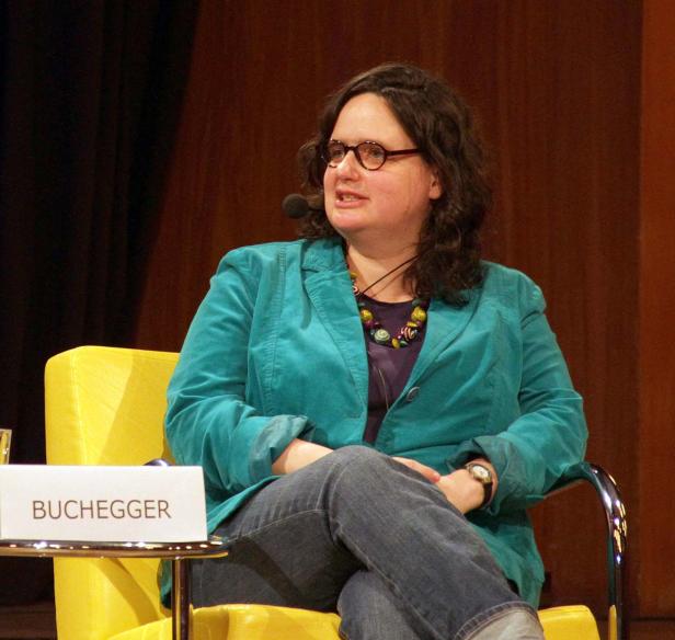 Eine Frau, vermutlich Buchegger, sitzt auf einem gelben Sessel und spricht in ein Mikrofon.