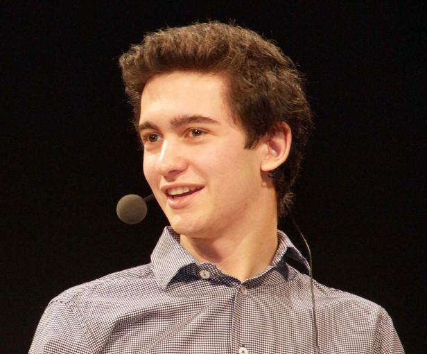 Ein junger Mann mit Mikrofon lächelt vor einem dunklen Hintergrund.