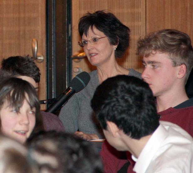 Eine Frau mit Brille spricht vor einer Gruppe von Zuhörern in ein Mikrofon.