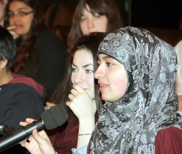 Eine junge Frau mit Hijab spricht in ein Mikrofon, während andere Personen im Hintergrund zu sehen sind.