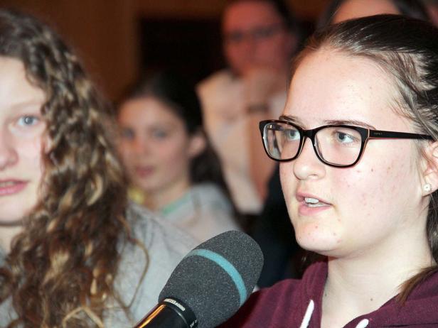 Ein junges Mädchen spricht in ein Mikrofon vor einer Gruppe von Zuhörern.