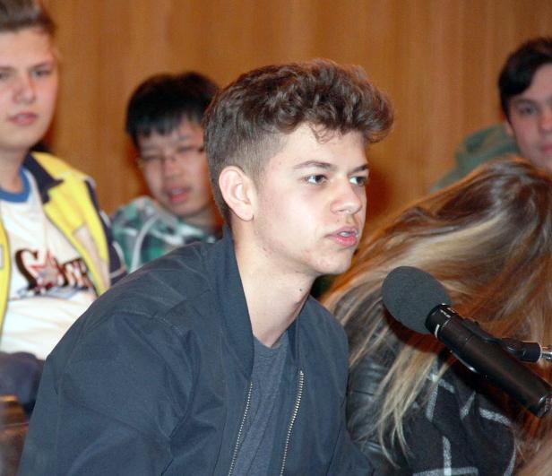 Ein junger Mann spricht in ein Mikrofon vor einer Gruppe von Zuhörern.