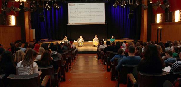 Publikum sitzt bei einer Podiumsdiskussion mit Johannes Bauer und anderen auf einer Bühne.