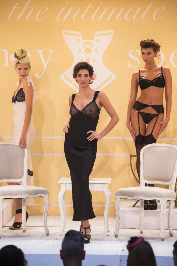 Drei Models präsentieren Dessous auf einer Bühne mit Stühlen.