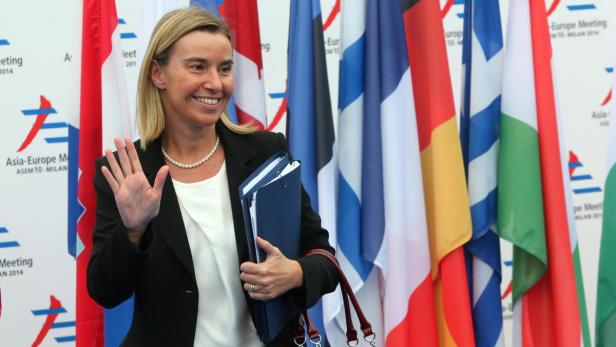 Eine Frau in einem schwarzen Blazer winkt vor einem Hintergrund von Länderflaggen.