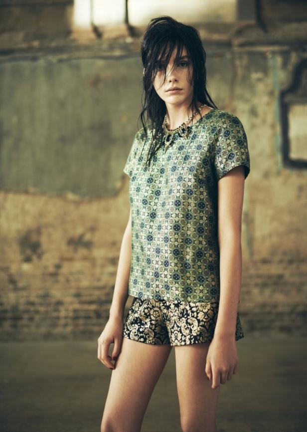 Eine junge Frau posiert in einem gemusterten Top und Shorts vor einer Wand.