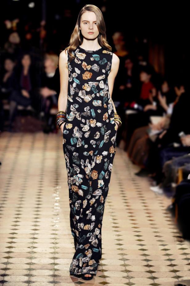 Ein Model präsentiert ein ärmelloses, langes Kleid mit einem Muster aus bunten Steinen auf einem Laufsteg.