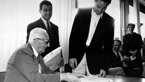 Enzo Ferrari unterschreibt ein Dokument, umgeben von anderen Personen.