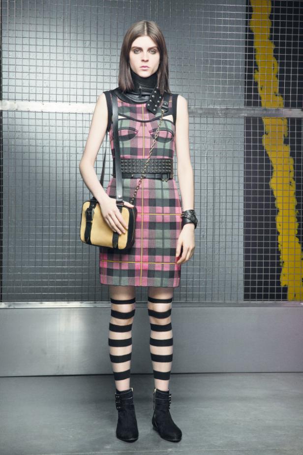Eine Frau in einem karierten Kleid mit Nietengürtel und gestreiften Strümpfen posiert vor einem Gitter.