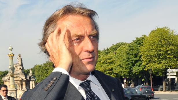 Ein Mann in Anzug steht vor einer Parkanlage und berührt seine Stirn.