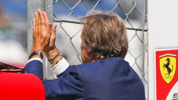 Eine Person im blauen Anzug klatscht hinter einem Zaun, neben einem Ferrari-Logo.