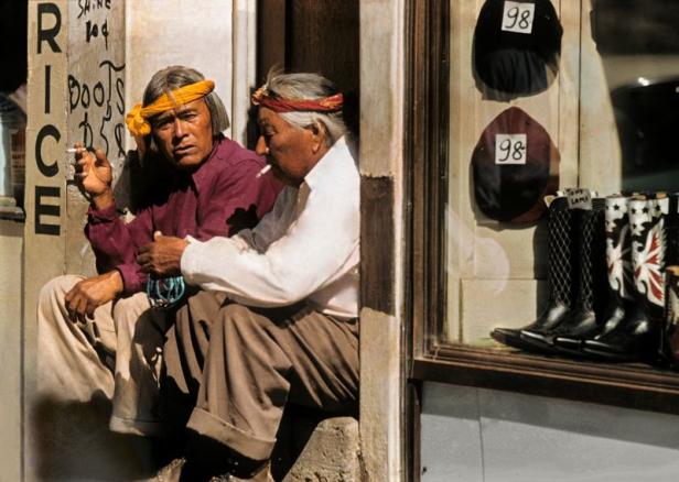 Zwei Männer mit Stirnbändern sitzen rauchend vor einem Laden mit Hüten und Stiefeln.