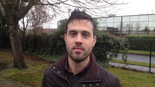 Ein Mann mit Bart steht im Freien vor einem Gebäude und Grünpflanzen.