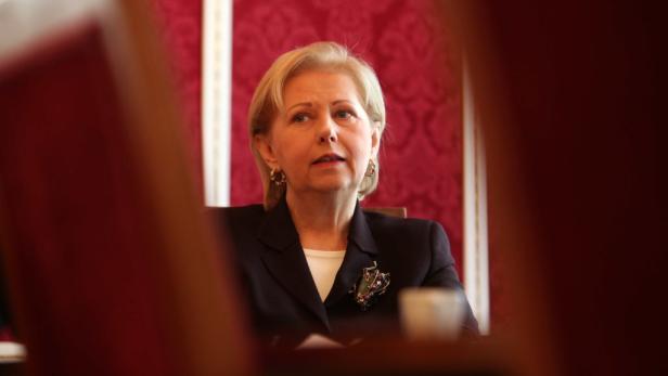 Eine Frau mit blonden Haaren und dunklem Blazer sitzt vor einem roten Hintergrund.