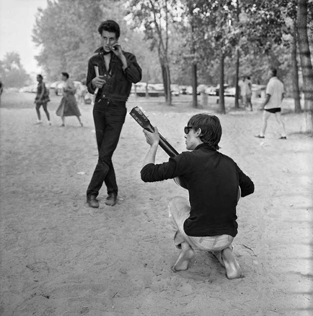 Ein Mann kniet barfuß im Park und spielt Gitarre, während ein anderer steht und zusieht.