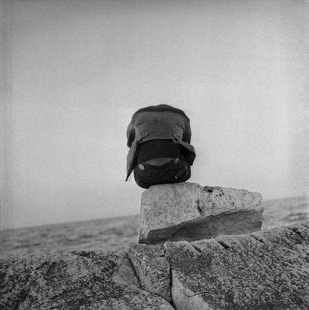 Eine Person sitzt auf einem Felsen am Meer, den Kopf gesenkt.
