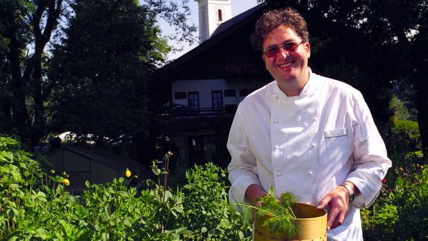 Ein lächelnder Koch mit Dill in einem Korb vor einem Haus mit Kirchturm.