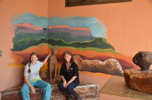 Geparden-Reportage Ein Mädchen und eine Frau sitzen vor einer Wandmalerei mit Leoparden und einer afrikanischen Landschaft.