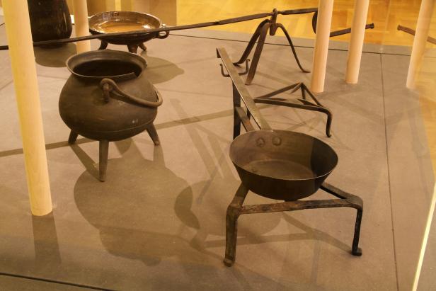 Verschiedene alte Kochutensilien aus Metall stehen auf einer Ausstellungsfläche.
