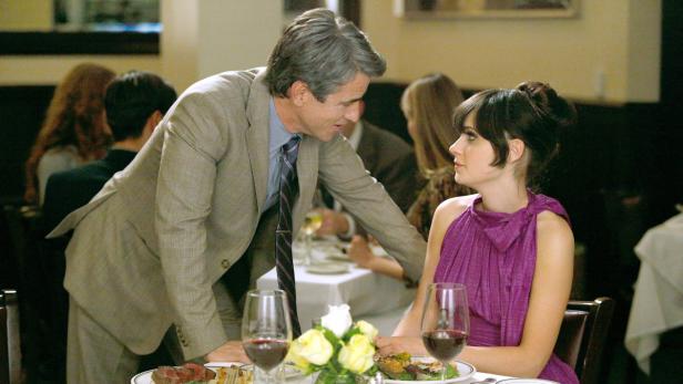 Eine Frau in einem lila Kleid sitzt an einem Tisch, während ein Mann sich zu ihr beugt.