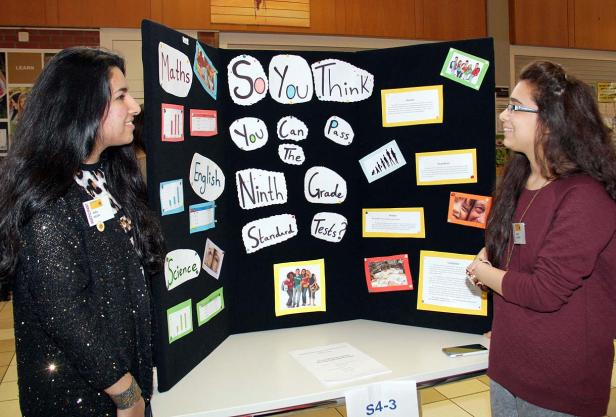 Zwei junge Frauen stehen vor einer Präsentation mit dem Titel „So You Think You Can Pass The Ninth Grade Standard Tests?“.