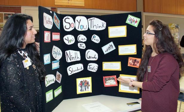 Zwei junge Frauen stehen vor einer Präsentation mit dem Titel „So You Think You Can Pass The Ninth Grade Standard Tests?“.