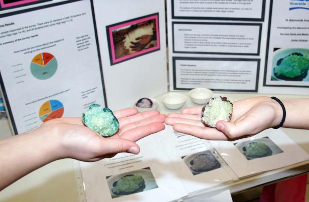 Zwei Hände präsentieren Kristalle vor einer Präsentationstafel über Diamantenabbau.