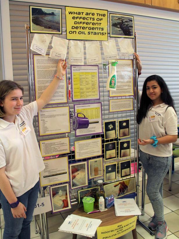 Zwei Schülerinnen präsentieren ein Projekt über die Wirkung verschiedener Reinigungsmittel auf Ölflecken.