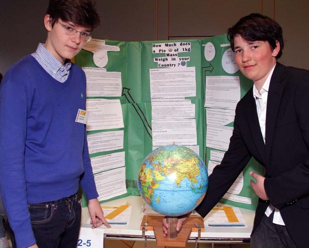 Zwei Jungen präsentieren ein Projekt über das Gewicht eines Kilos in verschiedenen Ländern, mit einem Globus und Infotafeln.