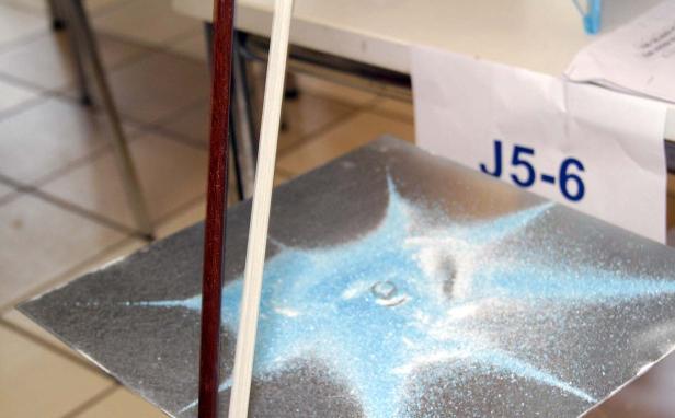 Ein Geigenbogen liegt neben einer Metallplatte mit blauem Sandmuster.