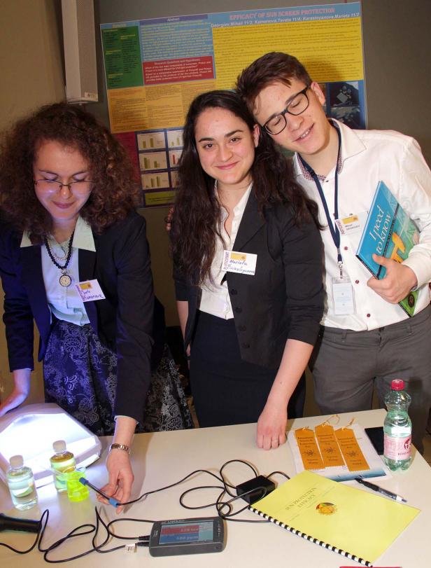 Drei junge Leute stehen an einem Tisch mit wissenschaftlichen Geräten und einem Poster zum Thema Sonnenschutz.