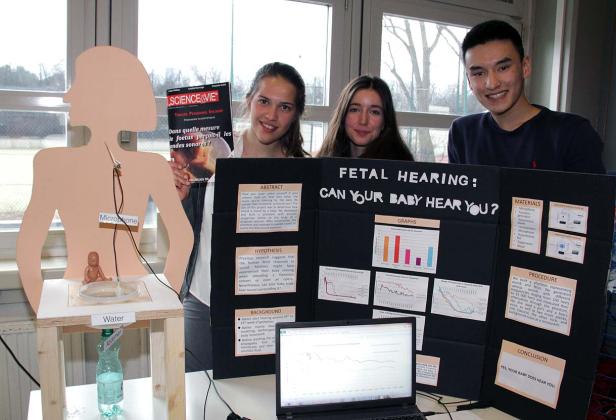 Drei Jugendliche präsentieren ein Projekt über fetales Hören mit Grafiken und einer Bauch-Attrappe.