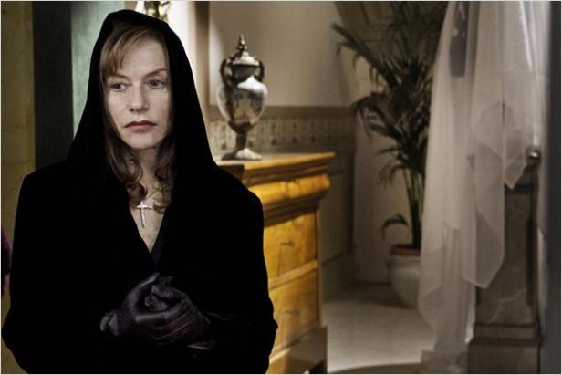 Isabelle Huppert trägt in einer Szene einen schwarzen Umhang und ein Kreuz.