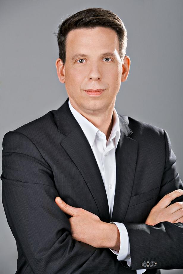 Ein Mann im Anzug mit verschränkten Armen vor grauem Hintergrund.