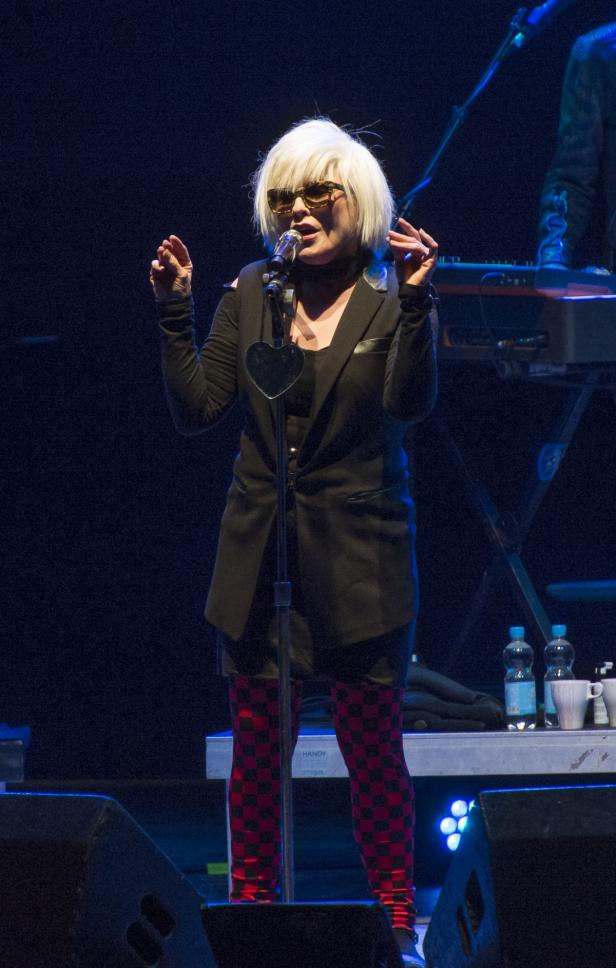Blondie, mit blonden Haaren und Sonnenbrille, singt auf der Bühne in ein Mikrofon.