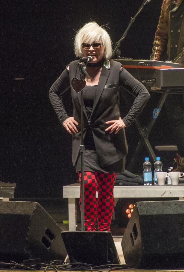 Blondie, mit Sonnenbrille und rot-schwarz karierter Hose, steht auf einer Bühne am Mikrofon.