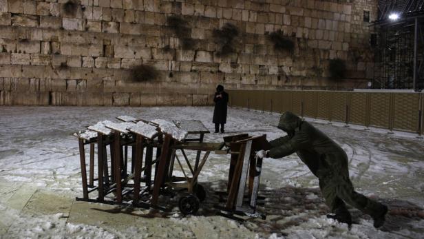 Eine Person schiebt einen Wagen mit Tischen vor der Westmauer in Jerusalem im Schnee.