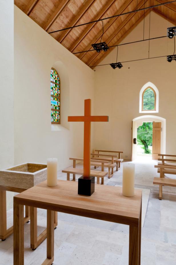 Innenansicht einer Kapelle mit Holzbänken, einem Kreuz und Kerzen auf einem Tisch.
