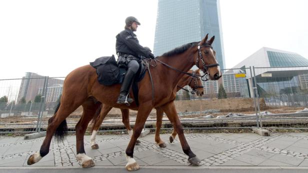 Ein berittener Polizist reitet auf einem braunen Pferd vor modernen Bürogebäuden.