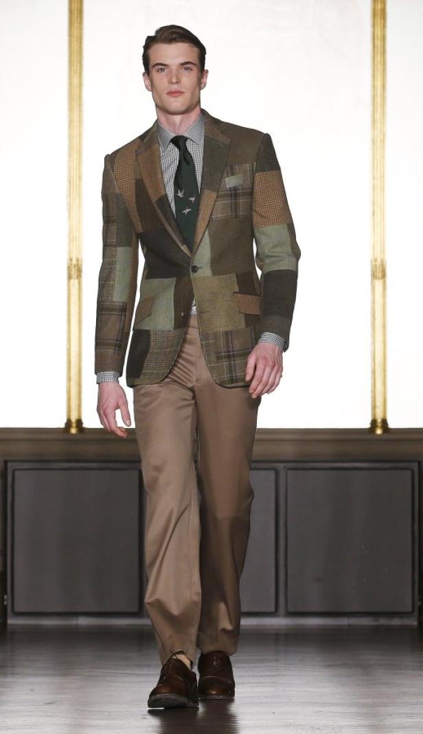 Ein Mann in einem Patchwork-Sakko, braunen Hosen und einer Krawatte mit Vogelmuster geht auf einem Laufsteg.