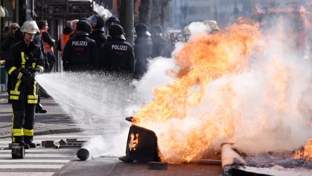 Ein Feuerwehrmann löscht einen brennenden Mülleimer, während Polizisten im Hintergrund stehen.