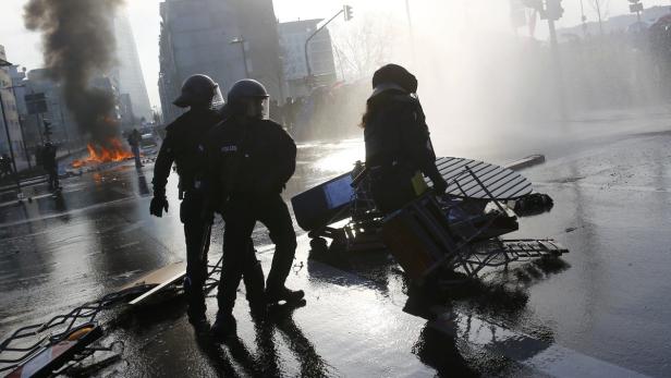 Polizisten stehen auf einer Straße, während im Hintergrund ein Feuer brennt und Wasserwerfer eingesetzt werden.