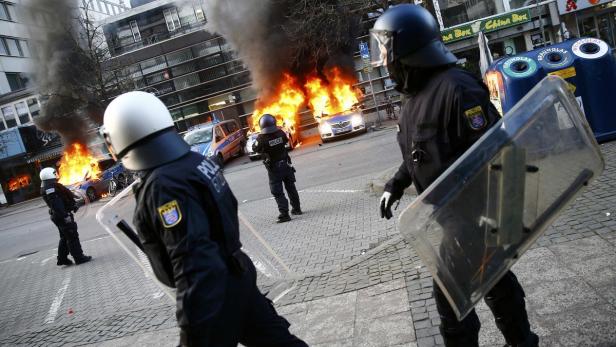 Polizisten mit Schilden stehen vor brennenden Autos auf einer Straße.